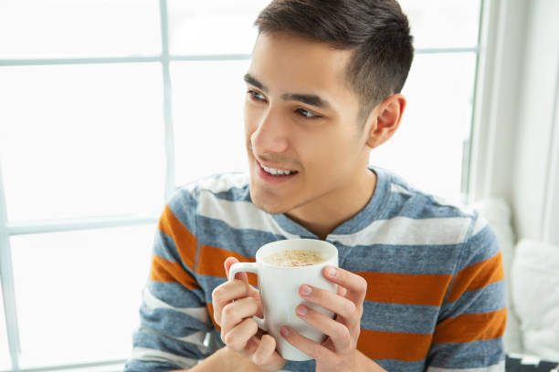 Casual smiling asian young adult drinking coffee up close at home.