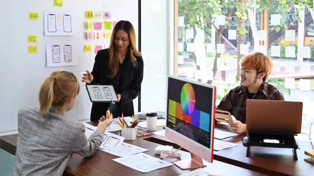 Photo of Female website designer showing user interface design on digital tablet to creative team at office presentation.