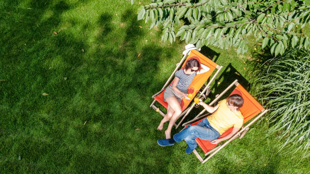 la pareja joven se relaja en el jardín de verano en tumbonas tumbonas sobre hierba, la mujer y los hombres toman bebidas en el picnic al aire libre en el parque verde el fin de semana, vista a�érea desde arriba - deck chair summer grass outdoor chair fotografías e imágenes de stock