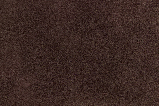 Background of dark brown suede matte fabric closeup. Velvet matt texture of umber nubuck textile. Velveteen pattern.