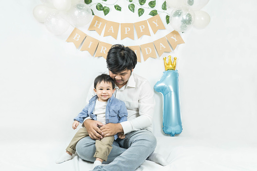 Asian baby boy celebrating first birthday,A father sits and hug his son smiling on a floor with a minimal background,white balloons and birthday flag.Selective focus