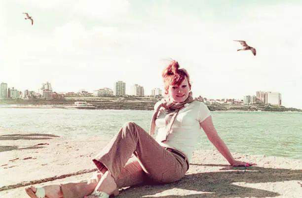 Vintage image of a woman sitting by the sea against a cityscape with seagulls flying around. Vintage image from the seventies.