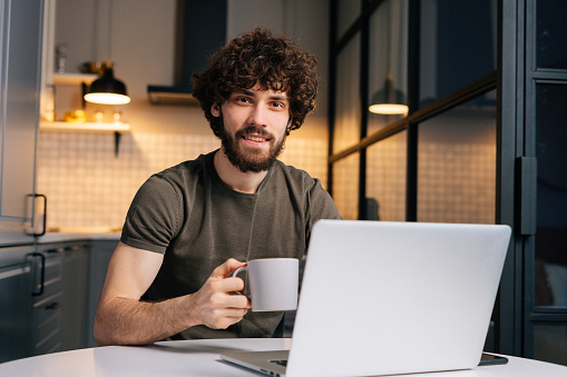 Portrait of smiling bearded young man holding in hand cup with morning coffee sitting at table with laptop computer, looking at camera, in kitchen with modern interior.
