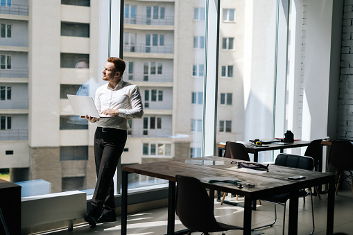 Portrait of thoughtful young business man working typing on laptop holding with one hand standing in modern office room near window, in sunny summer day on background of buildings, looking away.