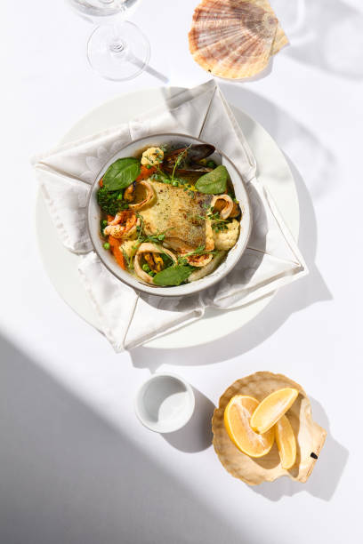 istock Elegant seafood stew with cod fish and vegetables in pot. Healthy fish dish - baked codfish, shrimp, mussels on broccoli and tomato. Serving white tablecloth and glass in summer day with shadow. 1392910012