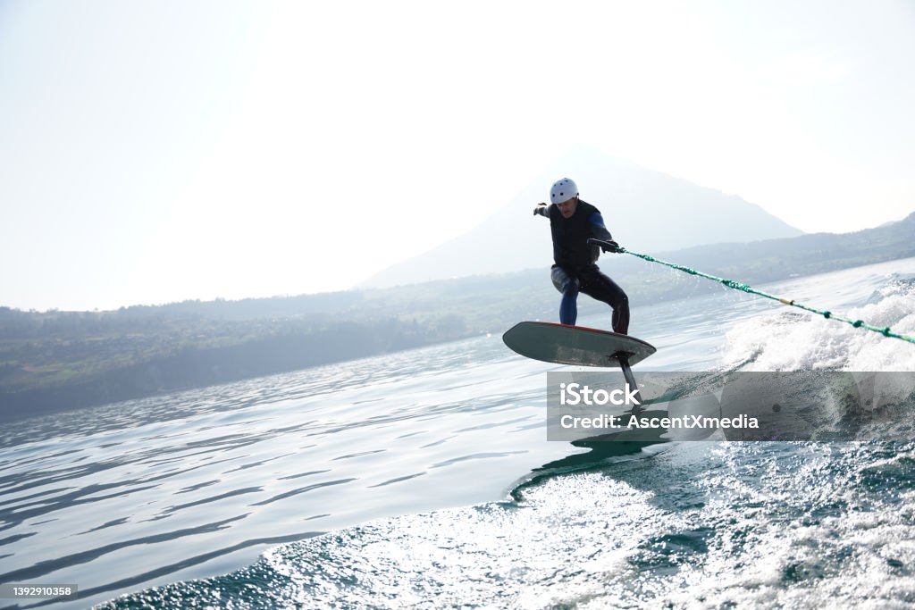 View of man riding wake foil behind motor boat Holding on to rope, he surfs the wake behind boat in Lake Thun, Swiss Alps behind Aquatic Sport Stock Photo