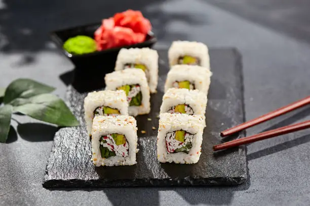 Maki sushi on dark stone table. Sushi roll california in sesame. Maki sushi with crab, avocado, cucumber inside, sesame outside. Style concept japanese menu with black background, leaves and hard shadow