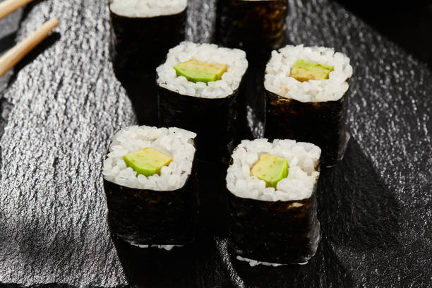 istock Avocado hosomaki on dark stone table. Hosomaki with avocado. Sushi roll with avocado inside. Style concept japanese menu with black background, leaves and hard shadow. 1392911889