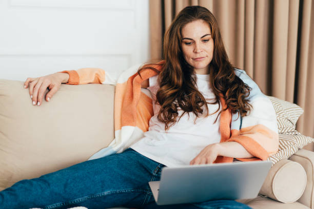 young concentrated business woman lying on sofa at home uses laptop to watch movie. - using laptop contemplation accessibility contemporary imagens e fotografias de stock