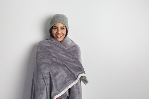 Happy Hispanic female warming with soft blanket and in beanie while standing against white background and looking at camera