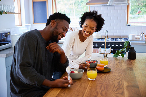 Young African couple talking and laughing together while eating a healthy breakfast at a kitchen island at home in the morning