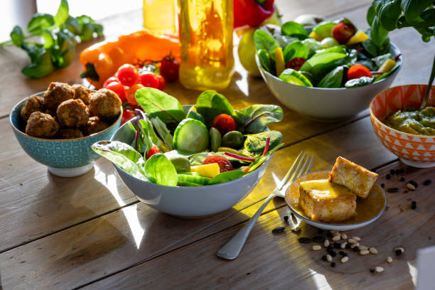 A close-up shot of a healthy vegan salad on a rustic wooden table, the ingredients are cherry tomatoes, spinach, olives, yellow pepper, cucumber, orange pepper, guacamole, falafel, and fried tofu.