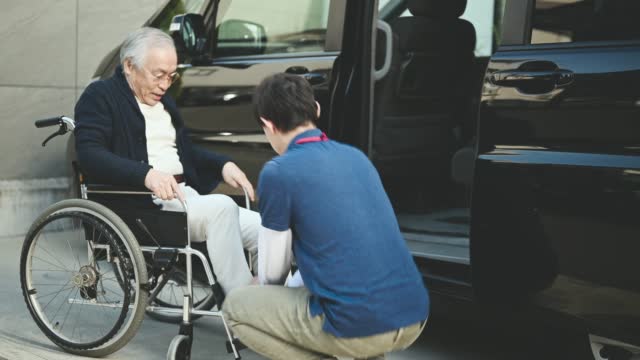 Young men caregiver helping senior man from car to wheelchair.