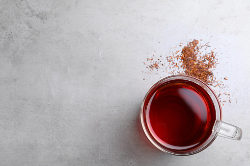 Freshly brewed rooibos tea and scattered dry leaves on grey table, flat lay. Space for text