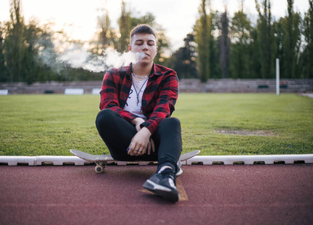 Teenage boy sitting down on skateboard and smoking stock photo