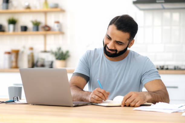 istock Positive modern Indian man, freelancer, designer or student, sitting at a kitchen at home, using laptop, studying or working online, smiling friendly, takes notes. E-learning concept 1392923470