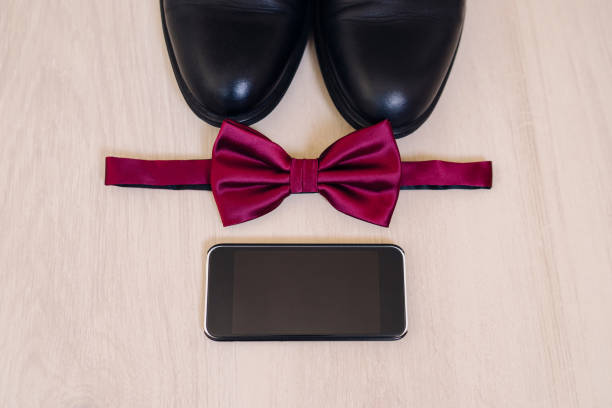 Top view of modern groom's outfit: leather shoes, bow tie, smartphone on wooden background. Flat lay composition.