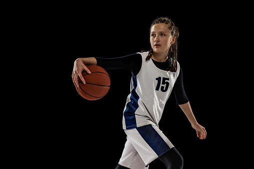 Training. Studio shot of teen girl in white uniform playing basketball, doing dribbling exercise isolated over black background. Concept of sport, active lifestyle, health, team game and ad