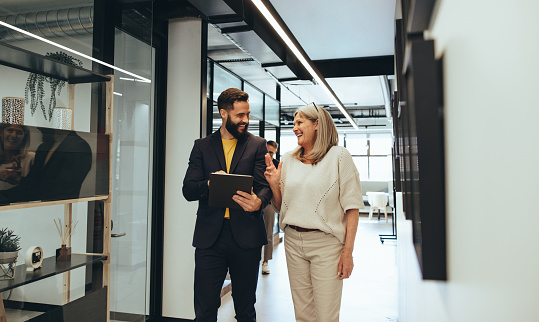 Diverse businesspeople sharing a laugh during a discussion in an office. Two happy business colleagues using a digital tablet while walking together in a modern workspace.