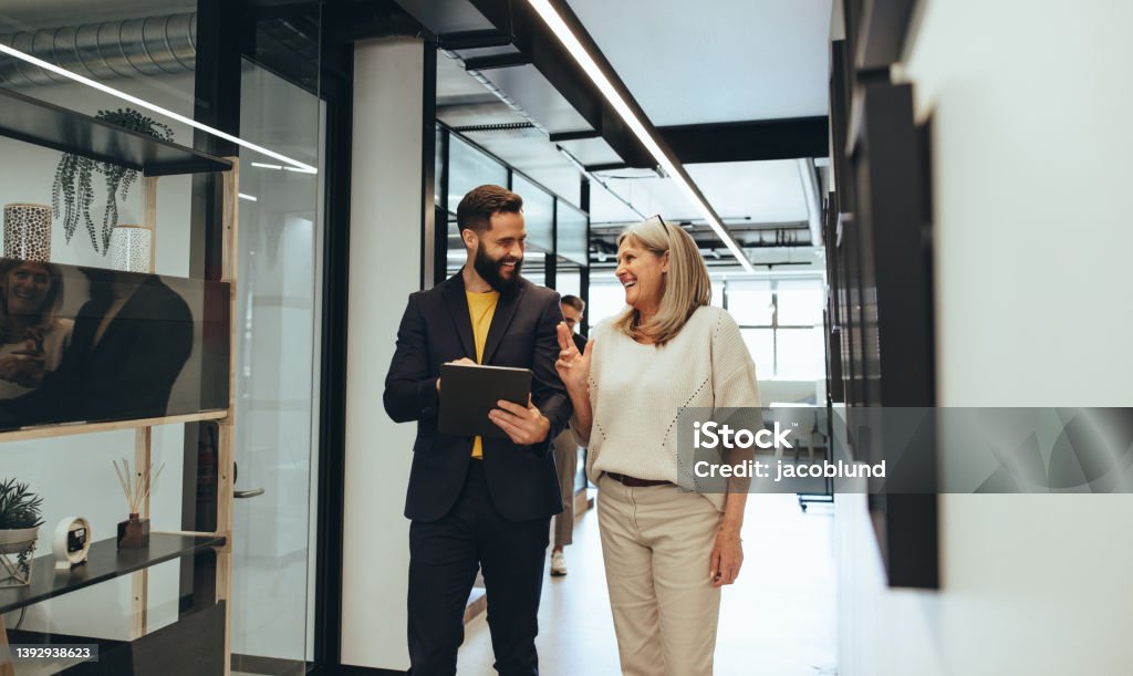 Diverse businesspeople laughing during a discussion Diverse businesspeople sharing a laugh during a discussion in an office. Two happy business colleagues using a digital tablet while walking together in a modern workspace. Office Stock Photo