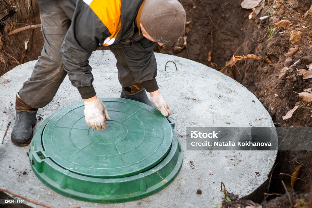 a utility worker lifts a manhole cover for sewerage maintenance and pumping out feces. Septic on a residential lot a utility worker lifts a manhole cover for sewerage maintenance and pumping out feces. Septic on a residential lot. Poisonous Stock Photo