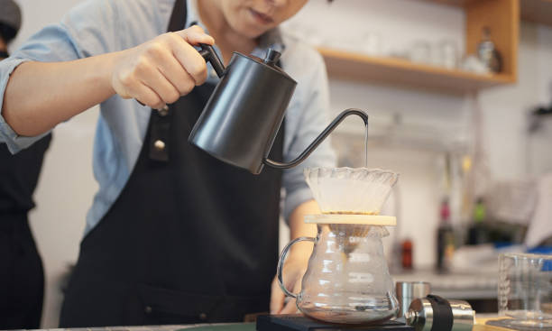 エプロンを着た女性カフェ経営者が、焙煎したコーヒーかすにお湯を注ぎ、店内のお客さんにコーヒーを用意。 - コーヒー　淹れる ストックフォトと画像