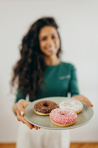 Portrait of the woman holding plate with different colour doughnut. She is wearing green blouse and khaki pants. She is wearing green blouse and khaki pants
