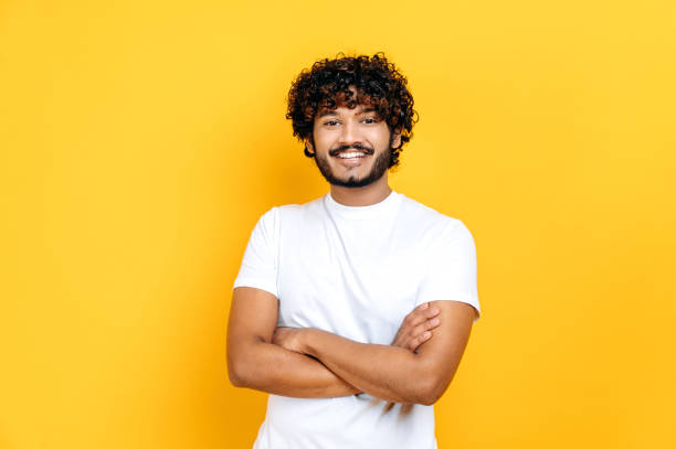 Joyful positive indian or arabian guy posing with arms crossed. Handsome millennial curly-haired man in basic white t-shirt stands on isolated orange background, looking at camera, smiling stock photo