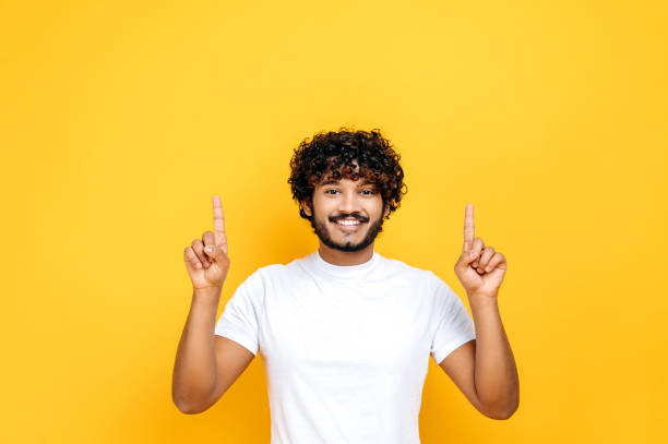istock Happy positive indian or arabian curly-haired guy in basic white t-shirt, pointing fingers up at empty mockup space for advertising, announcement, standing on isolated orange background, smiling 1392944436