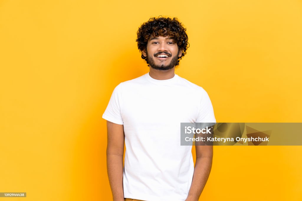 Portrait of handsome attractive positive curly- haired indian or arabian guy, wearing white basic t-shirt, standing over isolated orange background, looking at camera, smiling friendly Men Stock Photo
