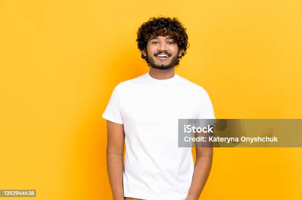 Portrait Of Handsome Attractive Positive Curly Haired Indian Or Arabian Guy Wearing White Basic Tshirt Standing Over Isolated Orange Background Looking At Camera Smiling Friendly Stock Photo - Download Image Now