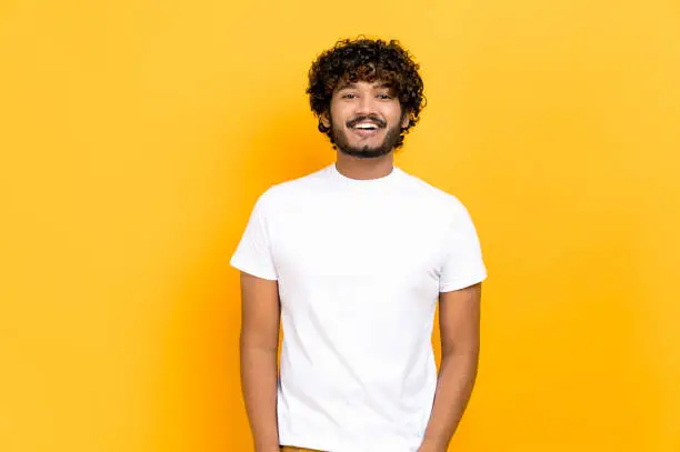 Photo of Portrait of handsome attractive positive curly- haired indian or arabian guy, wearing white basic t-shirt, standing over isolated orange background, looking at camera, smiling friendly