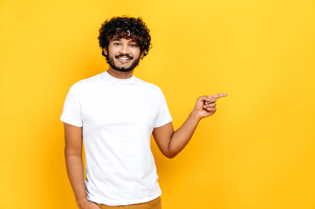 istock Happy curly haired millennial Indian young man, points with finger to empty space, looks at camera, smiling friendly, stands on isolated orange background in casual white t-shirt, mock-up concept 1392944439