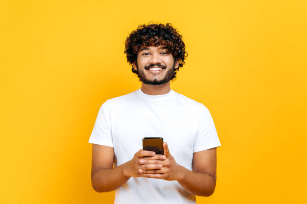 istock Handsome positive indian or arabian guy is using his smartphone, texting with friends, browsing the Internet and social networks, standing on isolated orange background, looks at camera, smiling 1392944443
