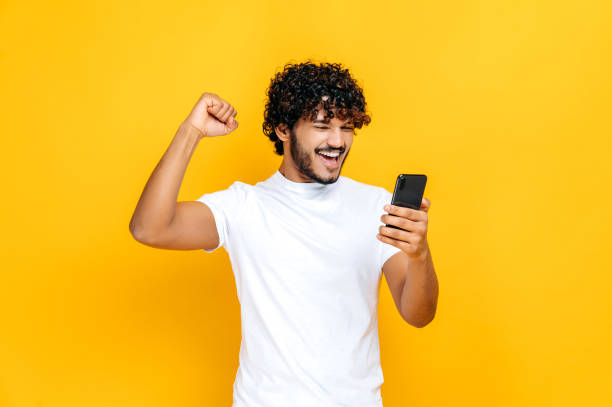 Amazed joyful excited Indian or Arabian guy holds smartphone, get unexpected news, winning lottery, stands on isolated orange background, cheerful facial expression, toothy smile, gesturing with fist stock photo