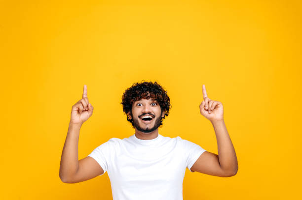 istock Happy indian or arabian guy in basic white t-shirt, amazed looks at the camera and points fingers up, at empty space, stands on isolated orange color background. Mock-up concept 1392944476