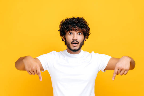 Positive indian or arabian happy guy in basic white t-shirt, amazed looks at camera and points fingers down, surprised face expression, stands on isolated orange color background stock photo