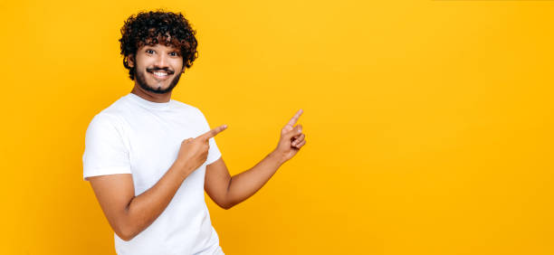 istock Amazed positive indian or arabian guy in basic white t-shirt, looks at the camera and points fingers to the side, at empty space, stands on isolated orange color background, smiling. Mock-up concept 1392944479