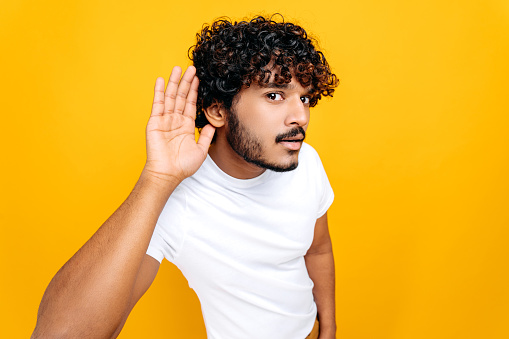 Deafness, gossip  concept. Positive Indian man, wearing basic t-shirt with hand near ear concentrated listening rumor, hearing gossip, standing on isolated orange background