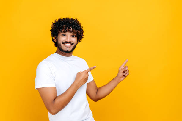 istock Excited happy indian or arabian guy in a basic white t-shirt, looks at the camera and points fingers to the side, at empty mock-up space, stands on isolated orange background, smiling. Mock-up concept 1392944483