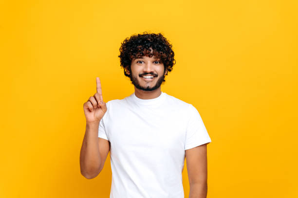 istock Joyful indian or arabian guy in basic white t-shirt, amazed looks at camera and points finger up, at empty copy space, stands on isolated orange color background, smiling. Mock-up, copy space concept 1392944489