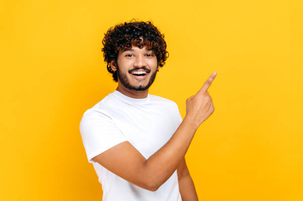 istock Cheerful indian or arabian guy in white basic t-shirt, looks and points finger to the side at mock-up space for your presentation, stands on isolated orange color background, looks at camera, smiling 1392944490