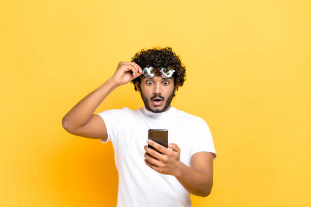 Amazed shocked unshaven Indian or Arabian guy holding smartphone in his hand, looking at the camera in surprise with his glasses raised, shocked facial expression, isolated orange background stock photo
