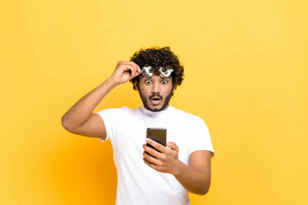 Photo of Amazed shocked unshaven Indian or Arabian guy holding smartphone in his hand, looking at the camera in surprise with his glasses raised, shocked facial expression, isolated orange background