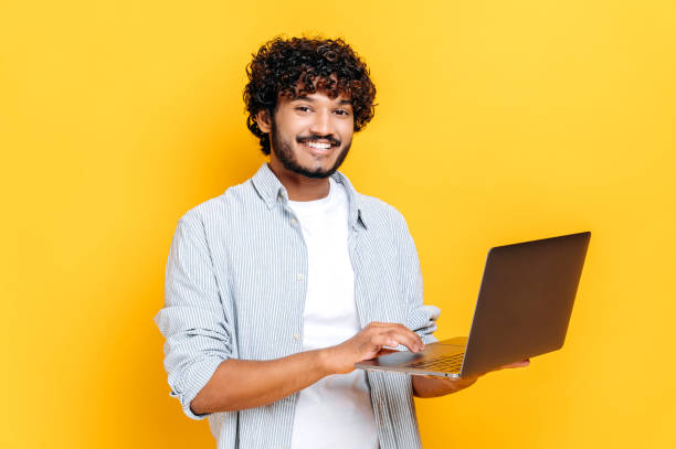 Attractive contented indian or arabian curly haired guy dressed in casual wear, holding an open laptop in his hand, looks friendly smiling at the camera, while standing on isolated orange background stock photo