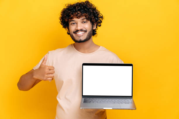 istock Excited cheerful Indian or Arabian guy in basic t-shirt, holds open laptop with blank white screen, showing thumbs up gesture, standing on isolated orange background, friendly smiling. Mock-up concept 1392944526