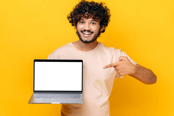istock Positive confident Indian stylish curly guy, holds an open laptop with blank white screen, points finger at it, standing on isolated orange background, smiling friendly. Copy space, mock-up concept 1392944533