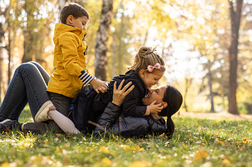 Happy young mother lying on grass, playing and bonding with her little daughter and son in park on sunny autumn day