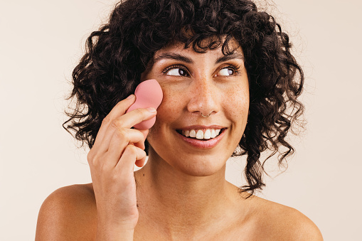 Pretty young woman looking away with a smile while using a makeup sponge on her face. Happy young woman using a beauty blender to apply makeup foundation on her face.