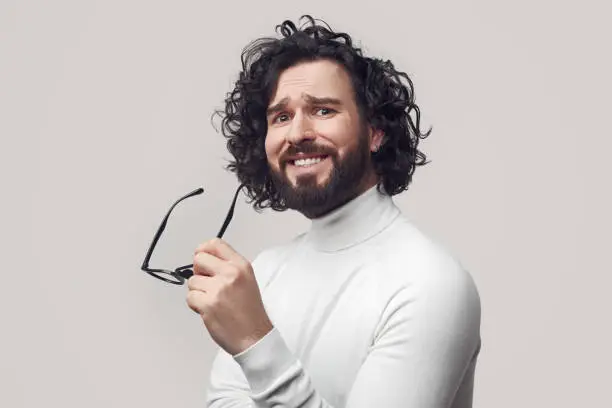 Young curly haired bearded guy with spectacles in hand looking at camera with astonished glance and doubt against gray background